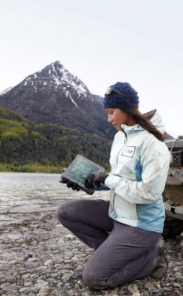 A woman in outdoor jacket and hat kneeling next to a river with a large mountain and forest in the background holding a Dell Latitude 12 Rugged Tablet (Model 7202) computer. A muddy vehicle can be seen in the background